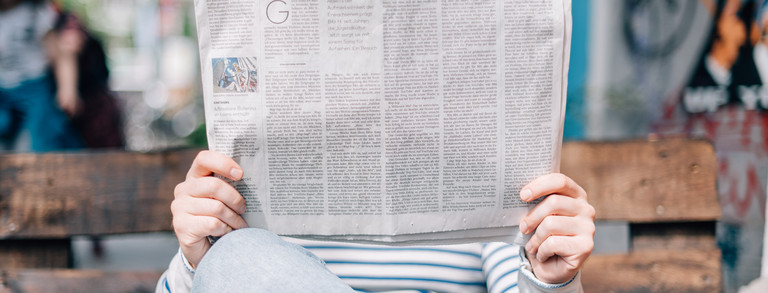 Man is sitting on a bench reading a newspaper.